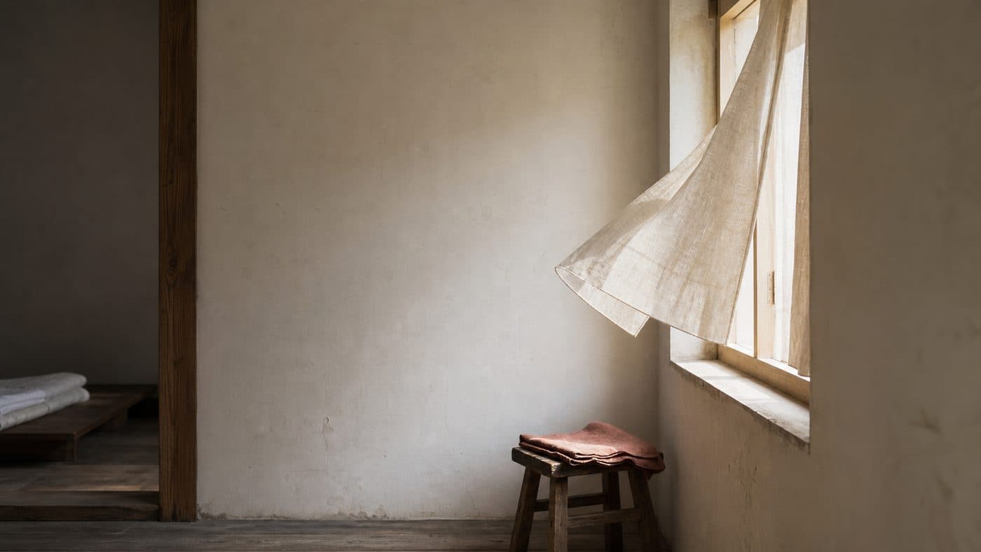 A plain room in morning light, a window open a hand-width with a light curtain lifting on the air.
